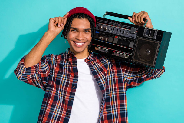Photo of positive funny optimistic guy with cornrows dressed checkered shirt hold boombox touching hat isolated on teal color background. - Fotó, kép