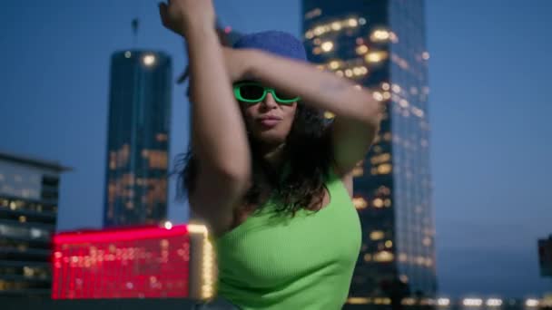 Actieve Afro-Amerikaanse zwarte vrouw in neon groene top en paarse emmer hoed dansen door stedelijke lichten achtergrond in het centrum 's nachts. Cool getalenteerde gemengde ras vrouwelijke danser beweegt ritmisch 4K Slowmo - Video