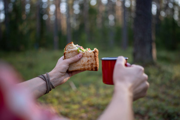 Man eating sandwich drinking tea having halt in forest enjoying nature, hands close-up. Guy hiker having break resting in woodland, mountains. Outdoors activity, camping, hiking, trekking concept. - Photo, Image