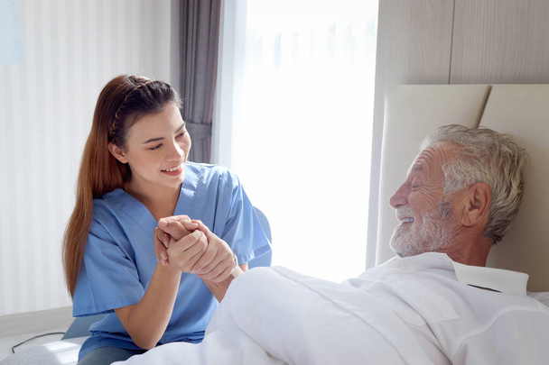 Усміхнений красивий догляд за жінкою-лікар тримає руку чоловіка старшого пацієнта, який лежить у лікарняному ліжку. Медсестра піклується про літнього чоловіка, який відчуває нудоту необхідність відпочивати в ліжку вдома, медична допомога літнім людям - Фото, зображення
