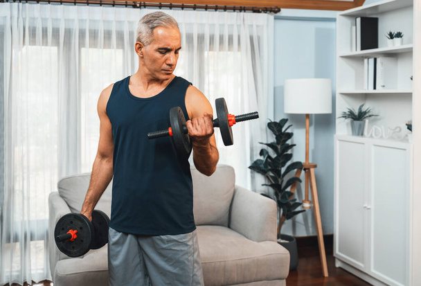 Athletic and sporty senior man engaging in body workout routine with lifting dumbbell at home as concept of healthy fit body with body weight lifestyle after retirement. Clout - Photo, Image