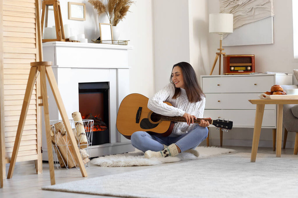 Молодая женщина играет на гитаре у камина дома - Фото, изображение