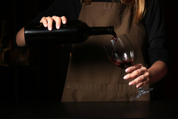 Γυναίκα μπάρμαν ρίχνει κόκκινο κρασί από μπουκάλι σε ποτήρι στο τραπέζι στο μπαρ - Φωτογραφία, εικόνα