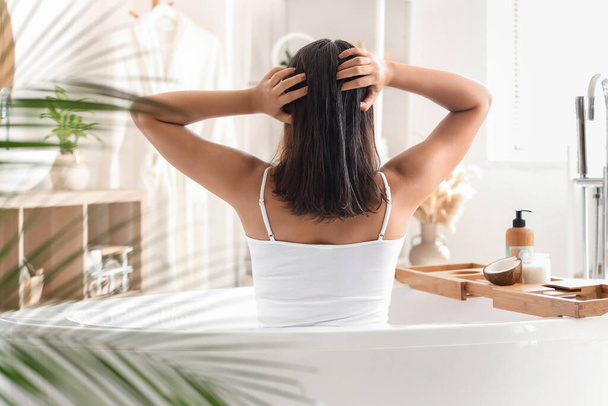 Jolie jeune femme appliquant de l'huile de coco sur ses cheveux dans la salle de bain, vue arrière - Photo, image