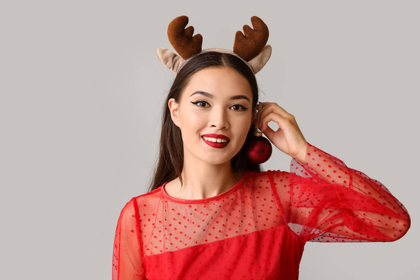 Hermosa joven asiática en cuernos de ciervo con bola de Navidad sobre fondo gris - Foto, imagen