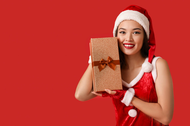Όμορφη νεαρή Ασιάτισσα γυναίκα ντυμένη σαν Άγιος Βασίλης με κουτί δώρου σε κόκκινο φόντο - Φωτογραφία, εικόνα