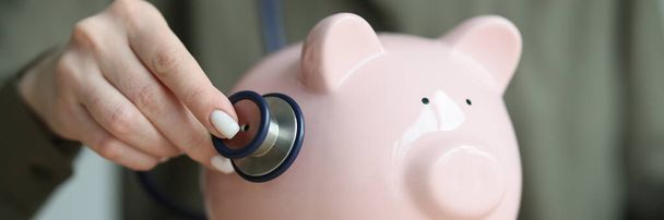 Woman checks condition of piggy bank with stethoscope sitting at wooden table. Medical equipment for checkup of pig for money savings - Photo, Image