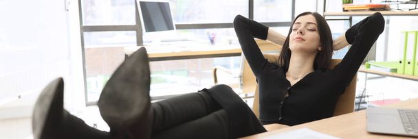Elegant brunette woman rests on chair putting legs in high-heels shoes on table in office. Young manager takes break form doing paperwork at workplace - Photo, Image