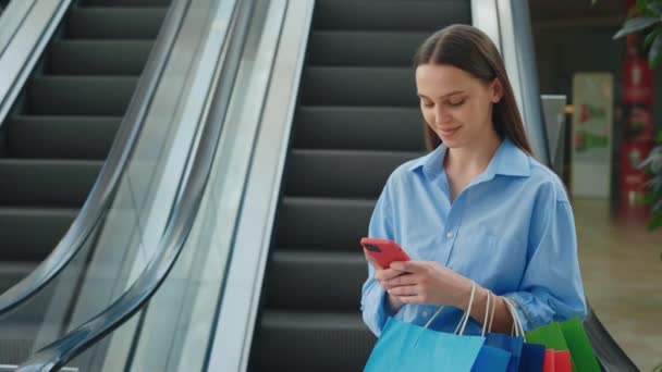 ハッピーな女性のお客様が紙袋に入れて購入しました. ショッピングバッグを持って 携帯電話を使って テキストメッセージを送ります。 エスカレーターの背景でオンラインショッピングにスマートフォンを使用している女性 - 映像、動画
