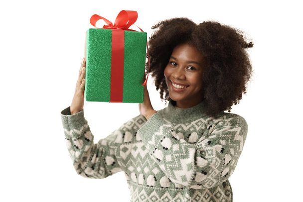 Junges schwarzes Mädchen im Strickpullover zeigt neben ihrem Gesicht lächelnd eine grüne Geschenkschachtel und blickt in die Kamera auf weißem Hintergrund - Foto, Bild