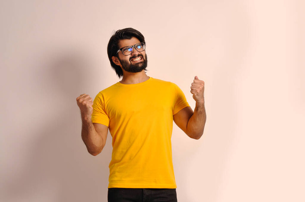Jeune homme barbu portant t-shirt et lunettes très heureux et excité de faire geste gagnant avec les bras levés, souriant et criant pour le succès. Fête - Photo, image