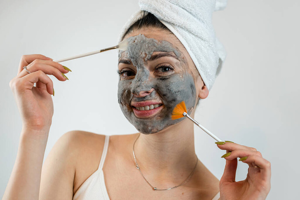 γυναίκα σε πετσέτα με βούρτσα εφάρμοσε μια μαύρη μάσκα καθαρισμού στο πρόσωπο ισοαλτωμένη σε λευκό. Αισθητική μάσκα ως αντιγηραντική θεραπεία - Φωτογραφία, εικόνα