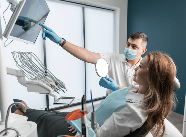 Modern technologies for installing dental implants allow patients to regain the natural appearance of a smile. Dentist showing screen to female patient in dental chair. High quality photo - Photo, Image
