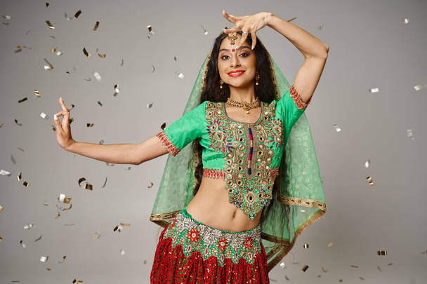 modelo feminino indiano atraente em roupas tradicionais posando em movimento cercado por confetes - Foto, Imagem