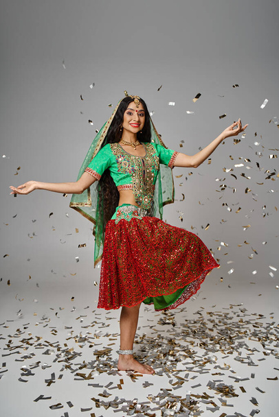 ελκυστική νεαρή ινδή γυναίκα σε πράσινο choli και κόκκινη φούστα ποζάρουν στο ένα πόδι κάτω από κομφετί βροχή - Φωτογραφία, εικόνα