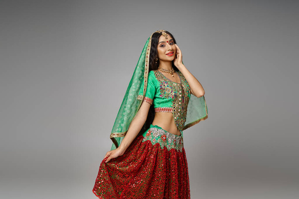 joyeuse femme indienne en choli vert touchant l'ourlet de sa jupe rouge posant avec la main sur la joue - Photo, image