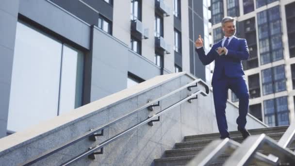 Iloinen, iloinen valkoihoinen liikemies tanssii kävellessään portaita modernin toimistorakennuksen taustalla, ulkopuolella. Hauska onnistunut liikemies puvussa ja juhlii voittoa ja menestystä - Materiaali, video