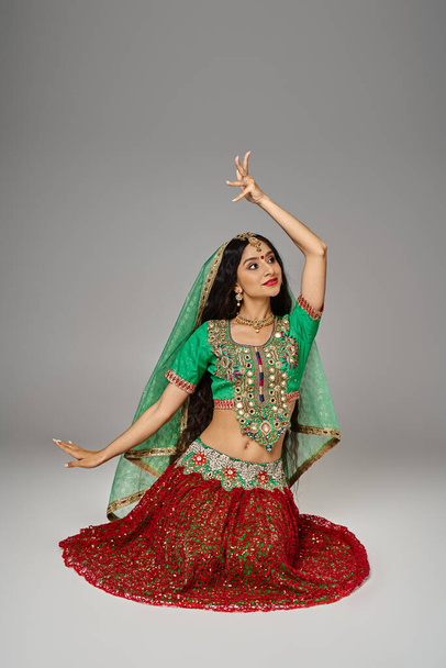 tiro vertical de mujer india joven en falda roja sentada en el suelo haciendo gestos y mirando hacia otro lado - Foto, Imagen