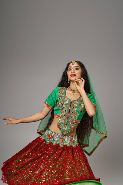 ελκυστική χαρούμενη Ινδή γυναίκα με bindi dot ποζάρει ενώ χορεύει ενεργά σε γκρι φόντο - Φωτογραφία, εικόνα