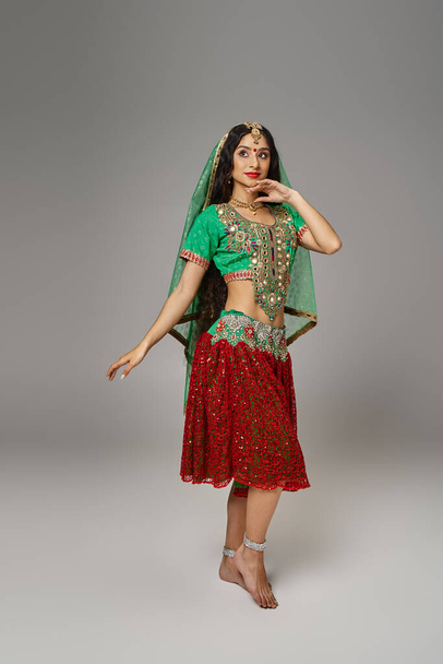 joven hermosa mujer india en verde choli y falda roja posando en movimiento sobre fondo gris - Foto, Imagen