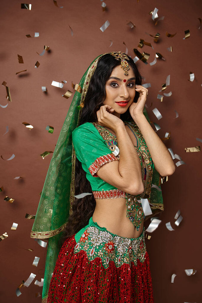 Ulusal kostümlü, konfeti yağmuru altında kameraya gülümseyen güzel bir Hintli kadının dikey çekimi. - Fotoğraf, Görsel