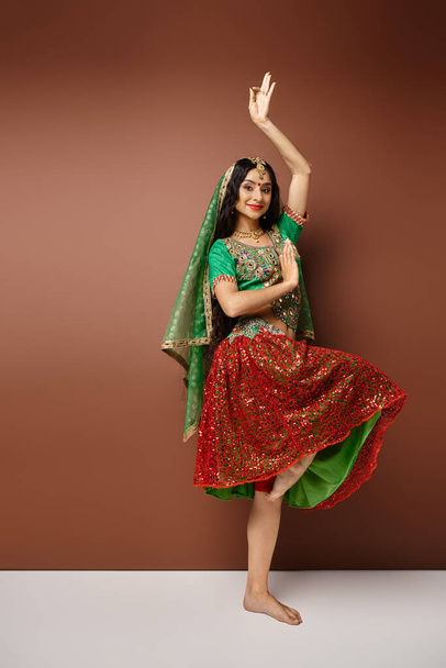 κάθετη λήψη της χαρούμενης νεαρής Ινδής με εθνική ενδυμασία που χορεύει ζωηρά και κοιτάζει την κάμερα - Φωτογραφία, εικόνα