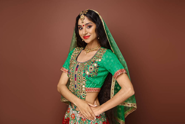 Yeşil peçeli ve bindi noktalı çekici Hintli kadın kahverengi arka planda kameraya gülümsüyor. - Fotoğraf, Görsel