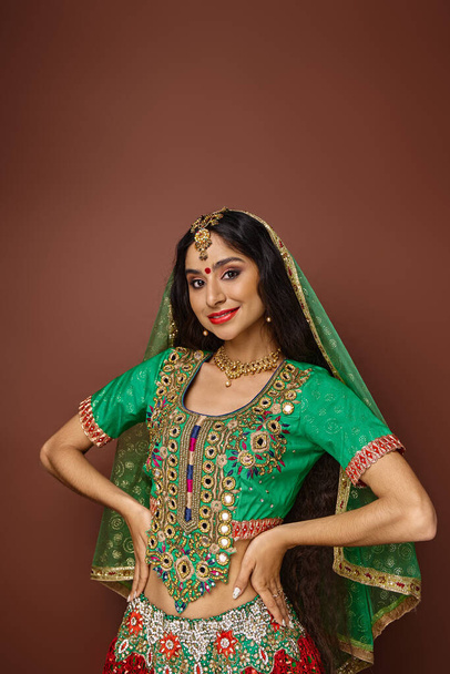 tiro vertical de jovem mulher indiana em roupas nacionais com braços akimbo sorrindo alegremente para a câmera - Foto, Imagem