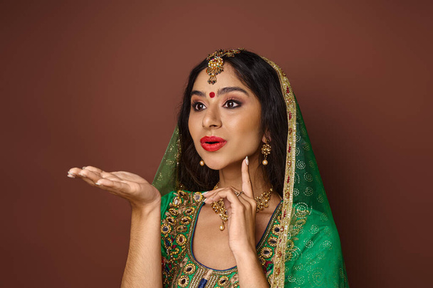 aantrekkelijke jonge indiaanse vrouw in groene sluier en bindi dot gebaren actief en kijken weg - Foto, afbeelding
