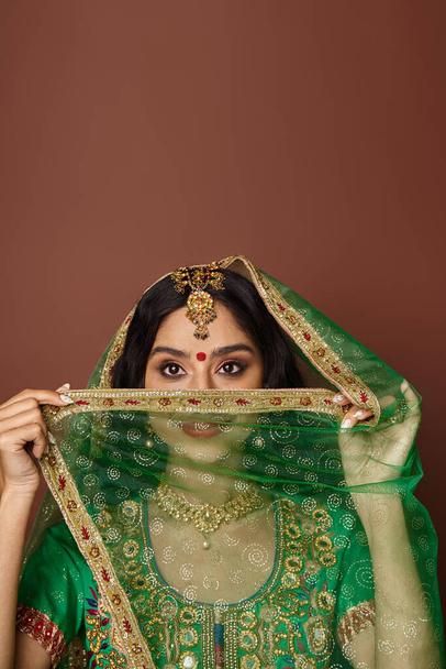 vertical shot of young indian woman in national costume with bindi covering her ace with green veil - Photo, Image