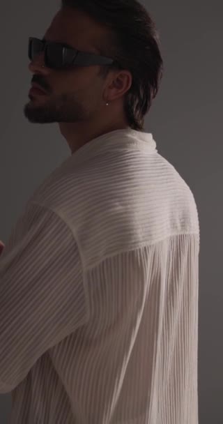 rear view of casual guy in white shirt rubbing palms, rubbing collar and adjusting sunglasses in front of grey background - Footage, Video