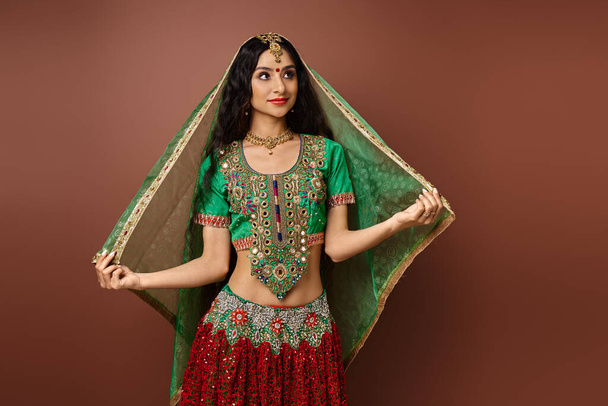 Ulusal kostümlü güzel Hintli kadın yeşil duvağa dokunuyor ve kahverengi arka plana bakıyor. - Fotoğraf, Görsel