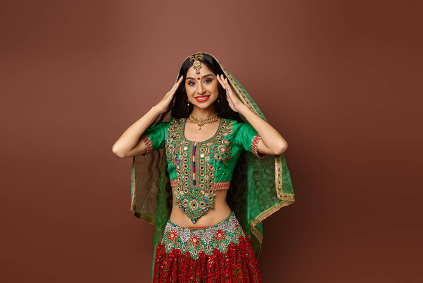 atractiva mujer india joven en ropa tradicional posando con las manos cerca de la cara sobre el telón de fondo marrón - Foto, imagen