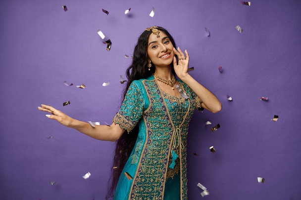 mujer india alegre con accesorios en sari azul sonriendo a la cámara posando bajo la lluvia confeti - Foto, Imagen