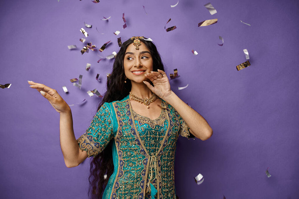 mulher indiana alegre em roupas nacionais com acessórios sorrindo e posando sob chuva confete - Foto, Imagem