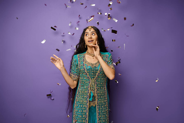 hermosa mujer india asombrada posando bajo la lluvia confeti sobre fondo púrpura con la mano cerca de la cara - Foto, imagen