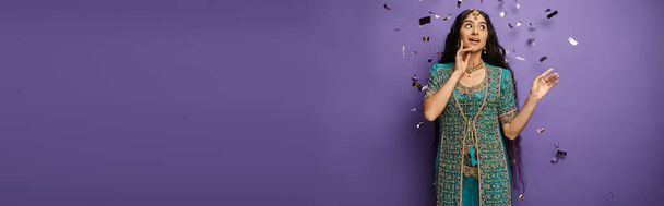 surprised young indian woman under confetti rain on purple backdrop with hand near face, banner - Photo, Image