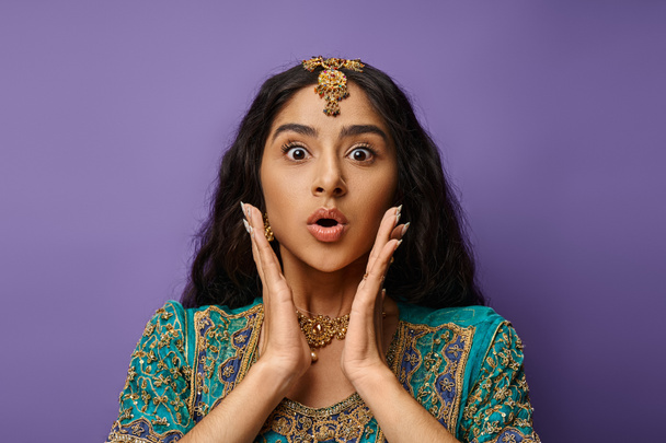 krásná šokovaná indická žena pózující na fialovém pozadí s rukama v blízkosti obličeje při pohledu na kameru - Fotografie, Obrázek