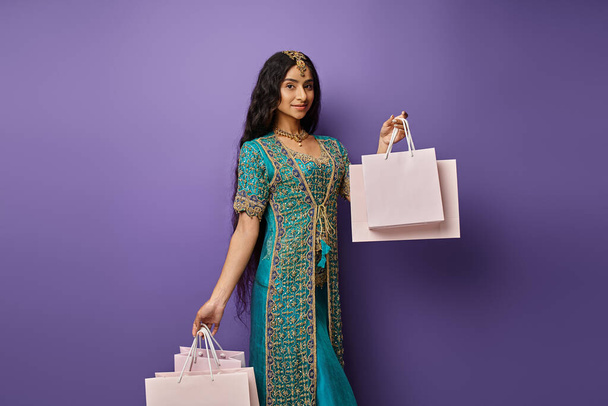 beautiful indian woman in blue sari with accessories holding shopping bags on purple backdrop - Photo, Image