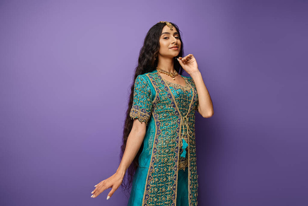 Mor arka planda eli yüzüne yakın poz veren mavi elbiseli neşeli Hintli kadın. - Fotoğraf, Görsel
