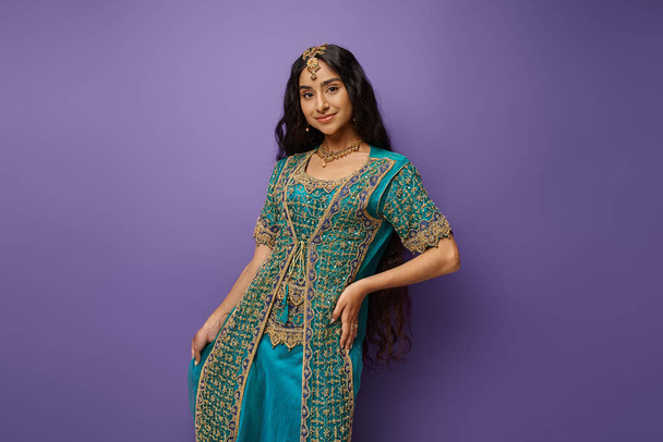 mujer india joven de buen aspecto en sari azul tradicional posando con la mano en la cadera sobre fondo púrpura - Foto, imagen