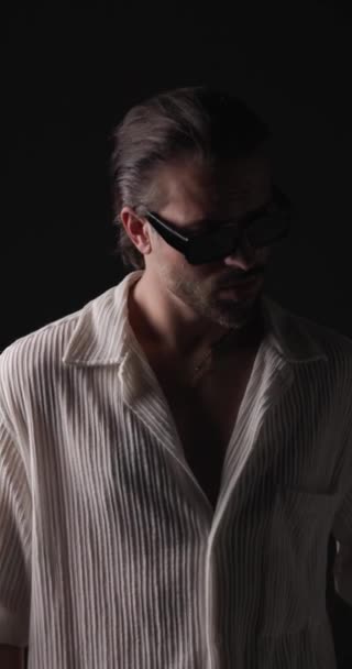 attraktiver, lässiger Mann mit Sonnenbrille, der zur Seite schaut, das Kinn berührt und nachdenklich ist, zur Seite schaut und vor Blitzlichtern auf grauem Hintergrund posiert - Filmmaterial, Video