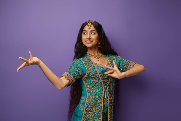 Веселая индийская женщина в традиционной одежде жестикулирует во время танцев и смотрит в сторону, фиолетовый фон - Фото, изображение