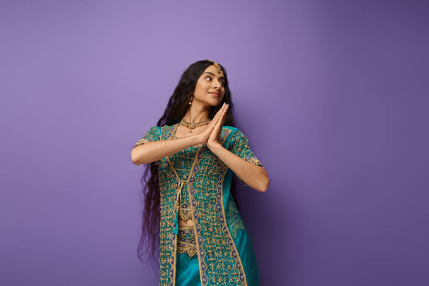 vrij vrolijke indische vrouw in blauw sari met accessoires poseren in beweging op paarse achtergrond - Foto, afbeelding