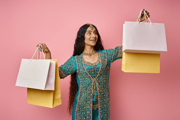 ピンクの背景にショッピングバッグを持っているアクセサリーと国民衣装の幸せなインドの女性 - 写真・画像