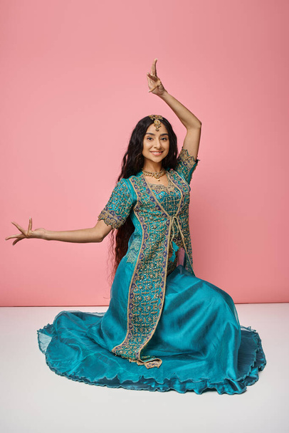 jolly indian woman in traditional blue sari sitting on floor and gesturing actively on pink backdrop - Photo, Image