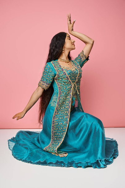 Ulusal kostüm giymiş genç Hintli kadın dans ederken pembe arka planda oturuyor. - Fotoğraf, Görsel