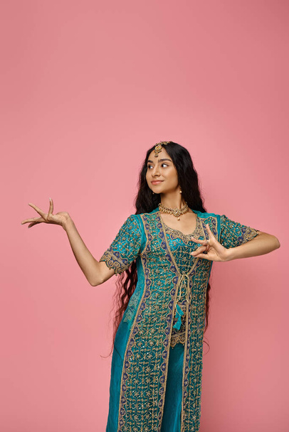 joyful attractive indian woman in traditional blue sari posing on pink background and looking away - Photo, Image