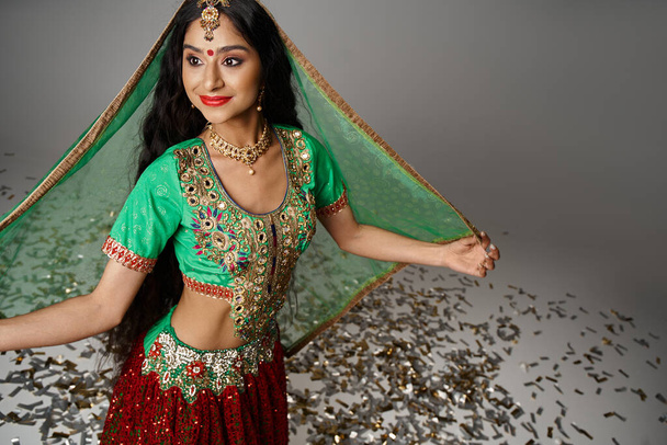 Радостная молодая индийская женщина в национальном костюме с точкой Бинди, позирующая на конфетти и отворачивающаяся - Фото, изображение