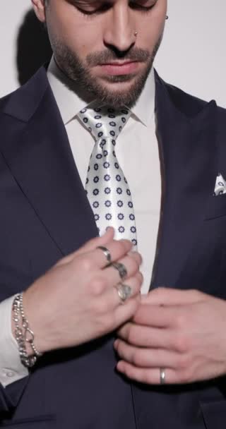 cool fashion man σε navy μπλε κοστούμι ρυθμίζοντας βραχιόλι, γραβάτα και κοστούμι, κοιτάζοντας προς τα πλάγια και με σιγουριά βάζοντας τα χέρια σε τσέπες σε γκρι φόντο - Πλάνα, βίντεο